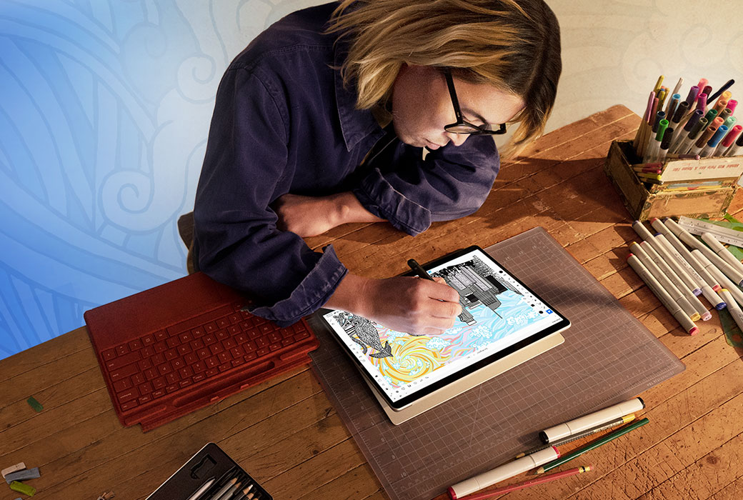 Artist and entrepreneur Daisuke Okamoto leans over a desktop to draw on a Surface Pro 8 tablet screen using a Surface pen. Also on the desktop are a detached Surface signature keyboard and drawing pencils.