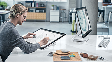 男人在 Surface Studio 螢幕上繪圖，一邊透過時尚辦公室中對面的另一台 Surface Studio 使用 Dial